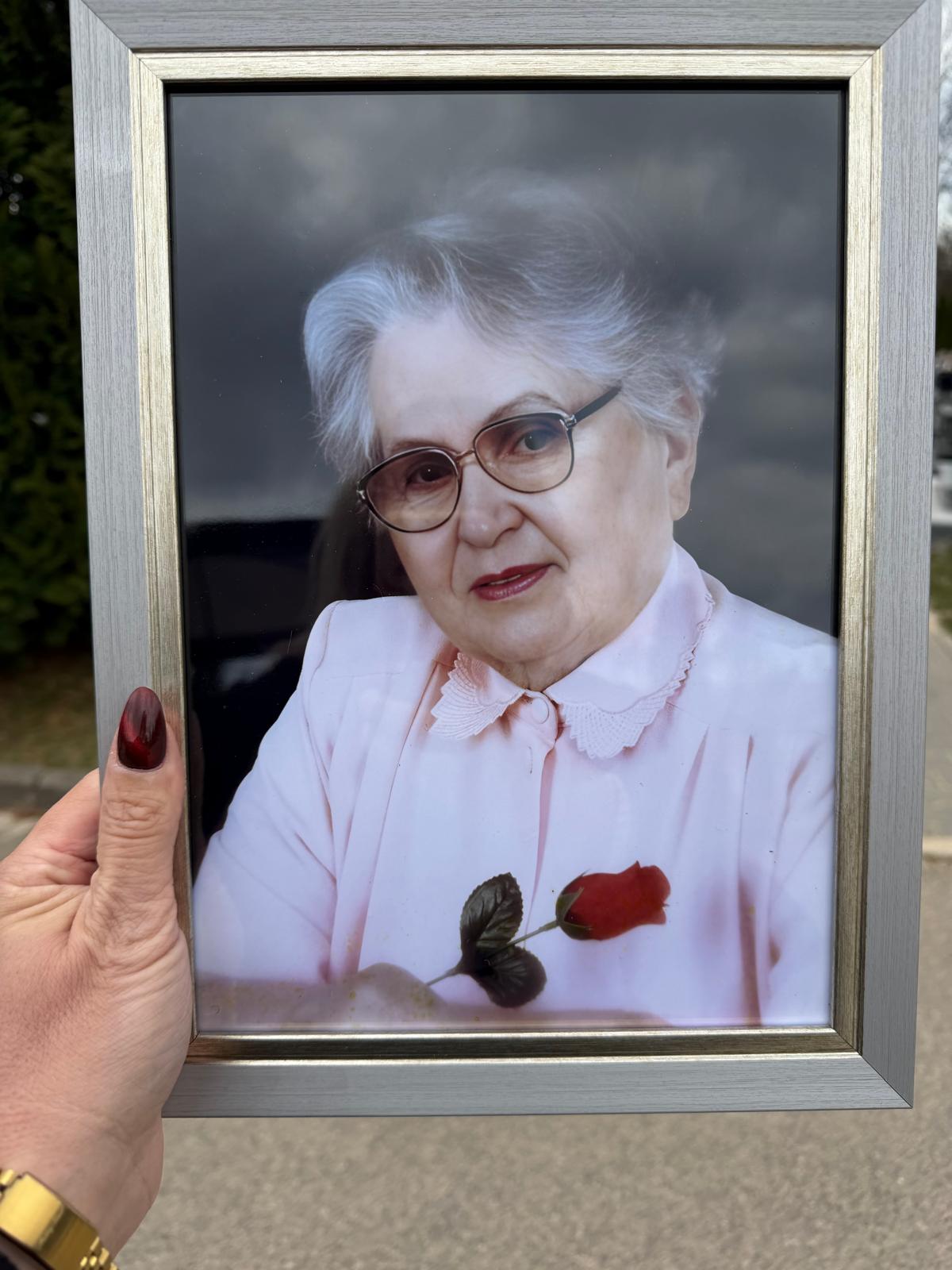 Ręka z bordowymi paznokciami trzymająca srebrną ramkę, w której znajduje się pamiątkowe zdjęcie starszej kobiety z siwymi włosami, w okularach, trzymającej czerwoną różę. W tle rozmyta droga i zieleń.