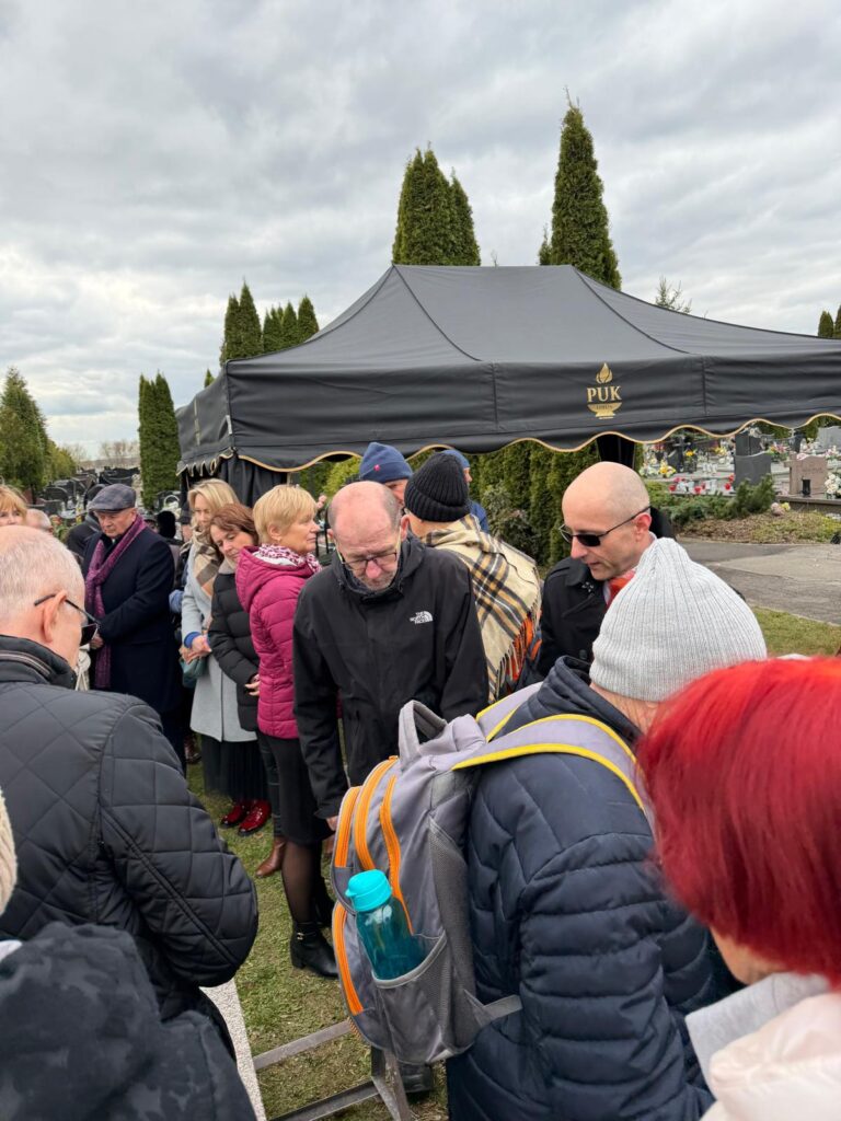 Grupa ludzi zgromadzona na cmentarzu podczas ceremonii pogrzebowej donatorów. W tle widoczny jest czarny namiot pogrzebowy oraz nagrobki.